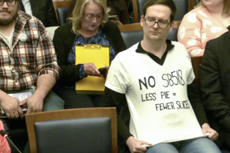 A man sits in a chair in the Indiana Statehouse wearing a homemade shirt that reads "No SB 518. Less pie equals fewer slices."