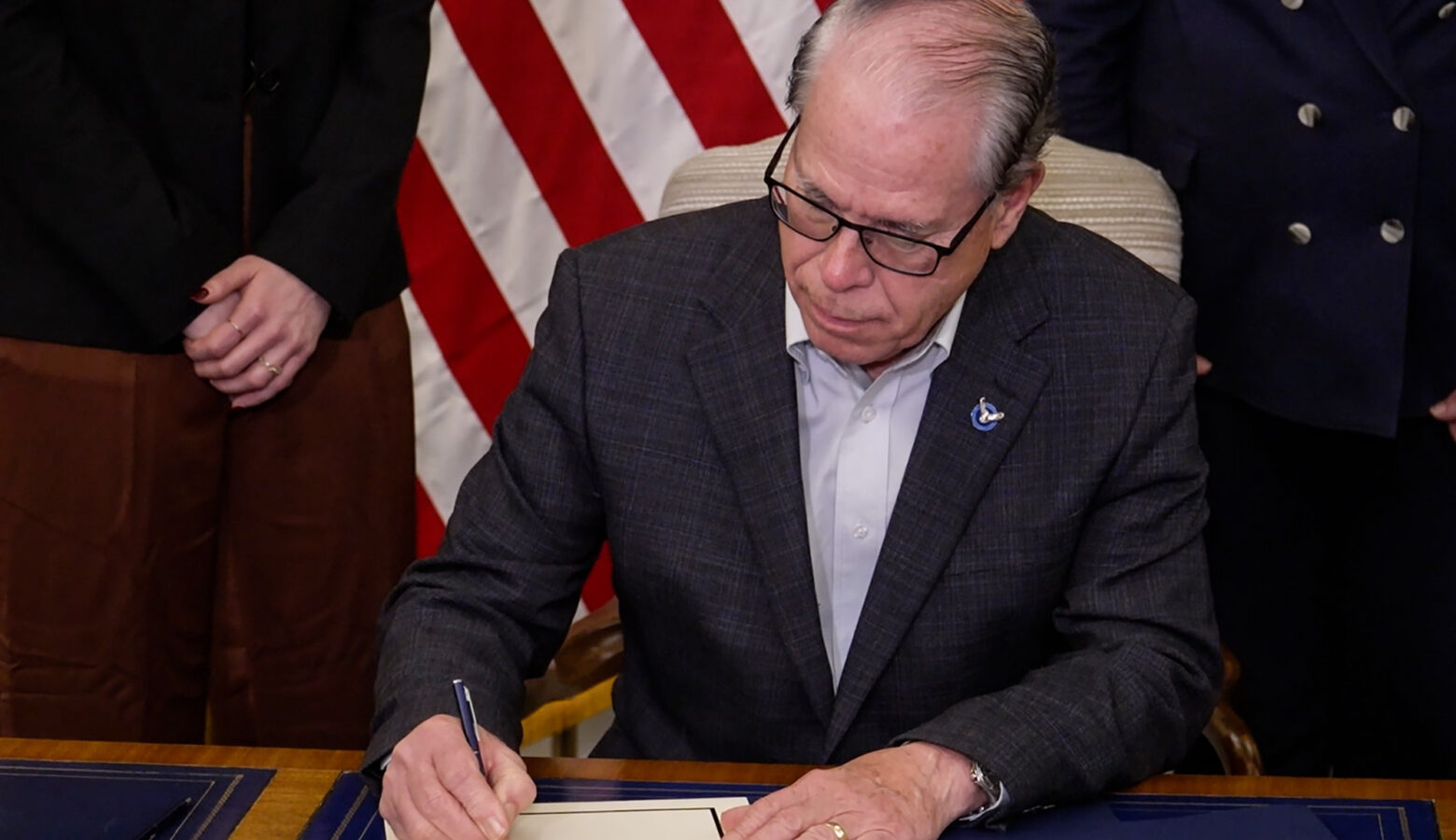 Governor Mike Braun signs one of the two executive orders he issued. He wears a plaid dark blue blazer with a white shirt underneath. He is a White man, bald and wears glasses. Behind him is an American flag that is flanked by two women whose faces aren't visible in the image.