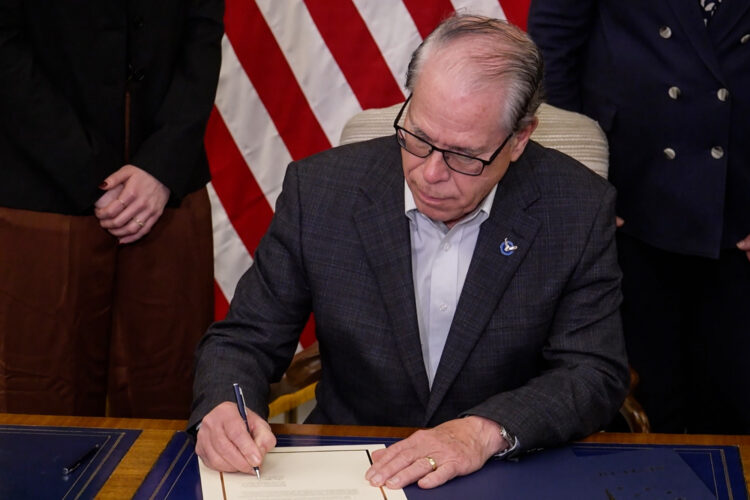 Governor Mike Braun signs one of the two executive orders he issued. He wears a plaid dark blue blazer with a white shirt underneath. He is a White man, bald and wears glasses. Behind him is an American flag that is flanked by two women whose faces aren't visible in the image.