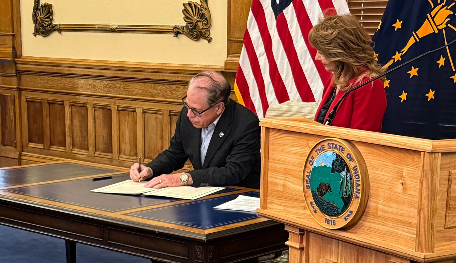 A wooden podium stands at the right side of a brown table. On top of the table are white pieces of paper. Governor Mike Braun wears a navy blue blazer and baby blue button up as he signs one of of the pieces of paper. Behind him are American and Indiana flags. His wife, Maureen Braun, stands to the right of him wearing a red blazer.