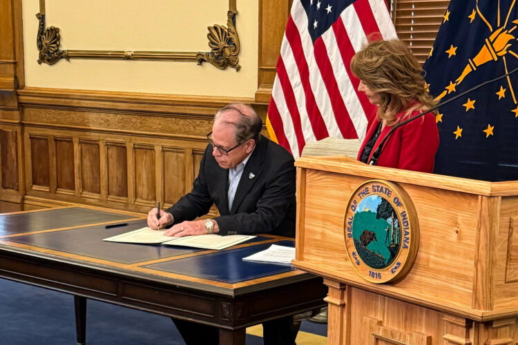 A wooden podium stands at the right side of a brown table. On top of the table are white pieces of paper. Governor Mike Braun wears a navy blue blazer and baby blue button up as he signs one of of the pieces of paper. Behind him are American and Indiana flags. His wife, Maureen Braun, stands to the right of him wearing a red blazer.