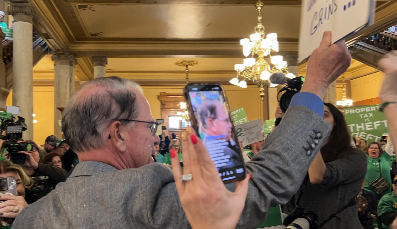 Mike Braun stands in the middle of a crowd of people, holding up a sign. Braun is a White man, balding with gray hair. He is wearing glasses and a light brownish-gray sportcoat.