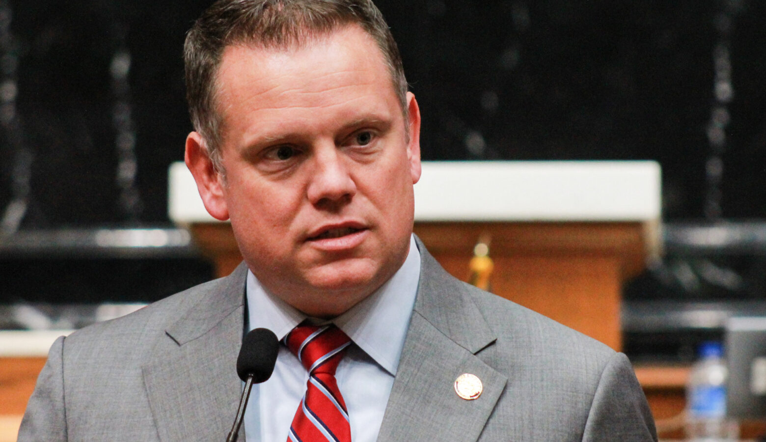 Chris Jeter speaks into a microphone on the House floor. Jeter is a White man with brown hair. He is wearing a gray suit and red striped tie.