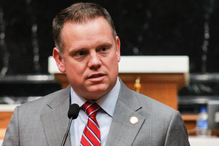 Chris Jeter speaks into a microphone on the House floor. Jeter is a White man with brown hair. He is wearing a gray suit and red striped tie.