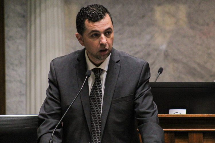 Fady Qaddoura wears a suit and leans forward to speak into a microphone.