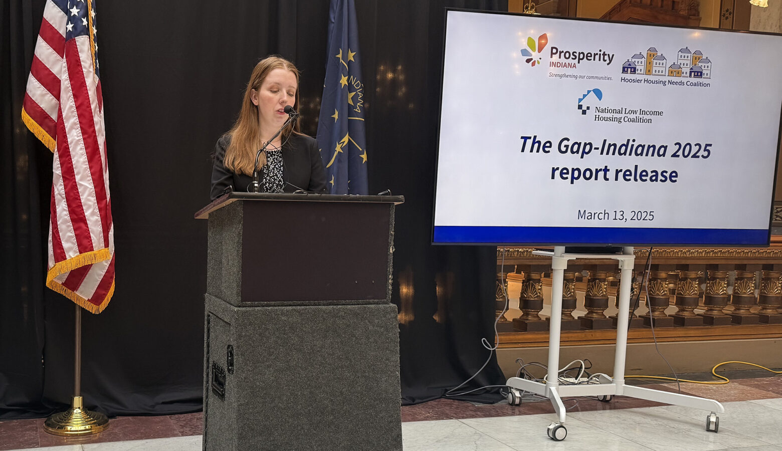 There is an American flag in front of a black curtain. There is a white woman with blonde hair speaking at a podium with a nearby screen that reads "The Gap-Indiana 2025 report release".