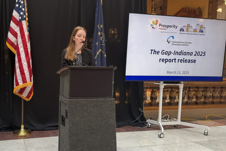 There is an American flag in front of a black curtain. There is a white woman with blonde hair speaking at a podium with a nearby screen that reads "The Gap-Indiana 2025 report release".