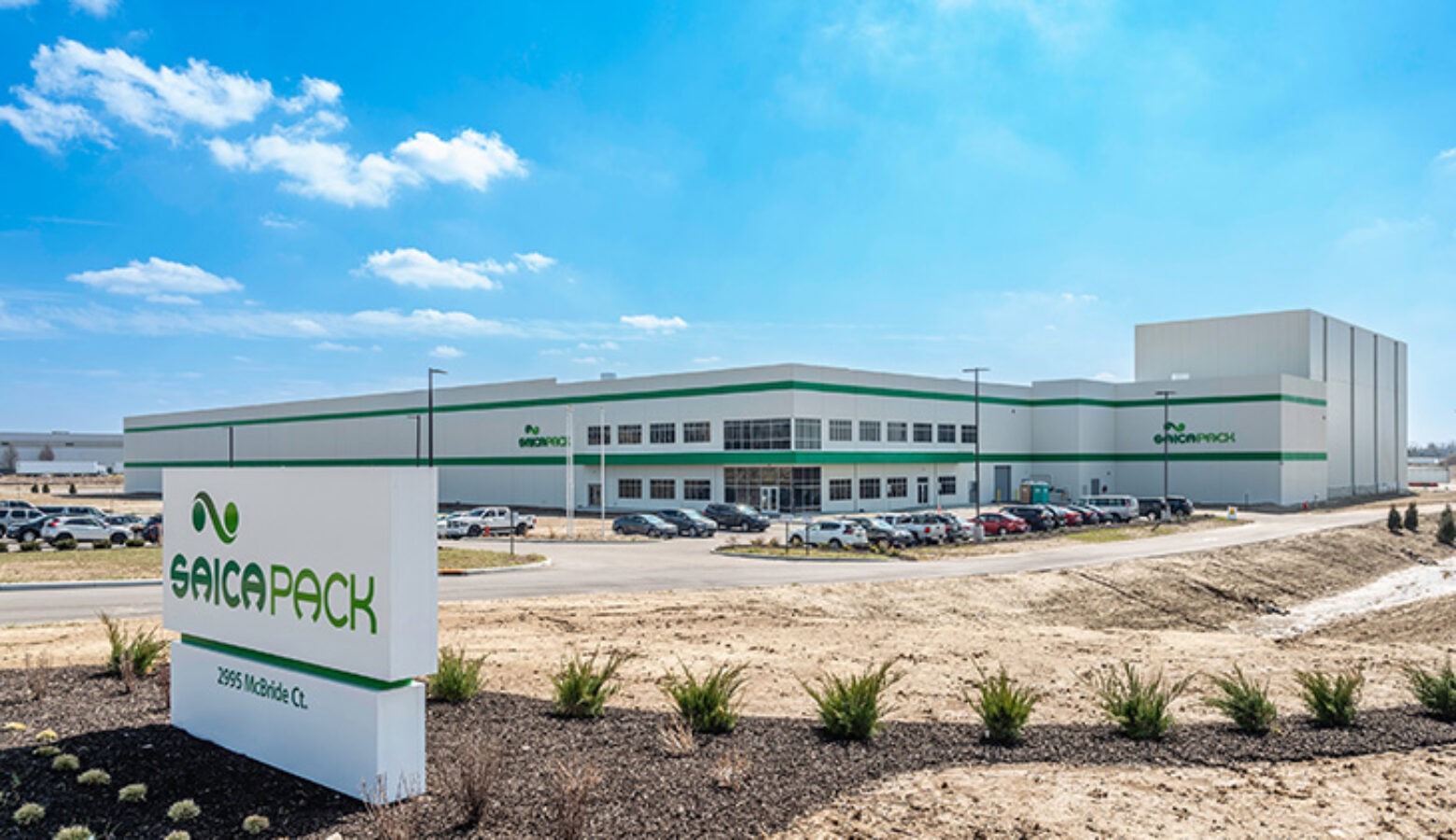 A while building with two green stripes. A parking lot filled with cars surrounds it. In front is a sign that says Saica Pack.