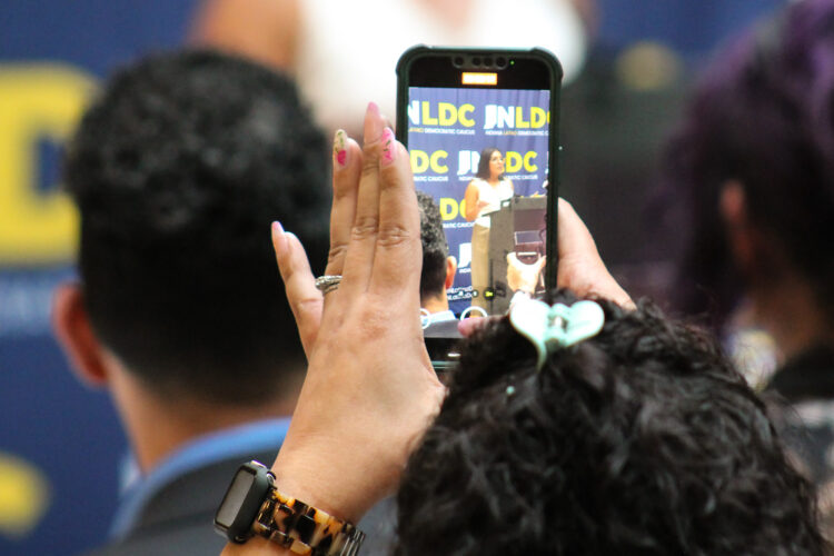 The Indiana Latino Democratic Caucus vice president speaks at a podium as a member of the audience records her with their phone. The audience member is facing away from the camera, but they have dark curly hair in a claw clip and a tortoiseshell watch band.