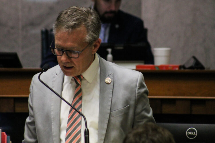 A man in a gray suit jacket with a gray and salmon striped tie looks down while speaking into a microphone.