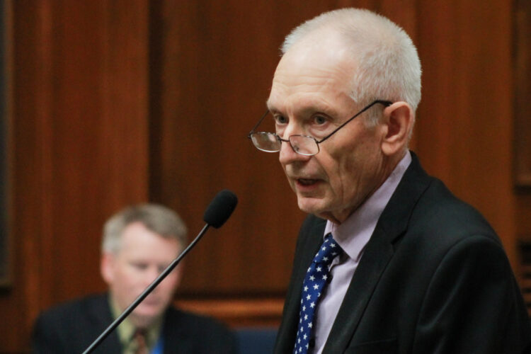 Jeff Thompson speaks into a microphone on the House floor. Thompson is a White man with white hair. He is wearing glasses and a suit and tie.