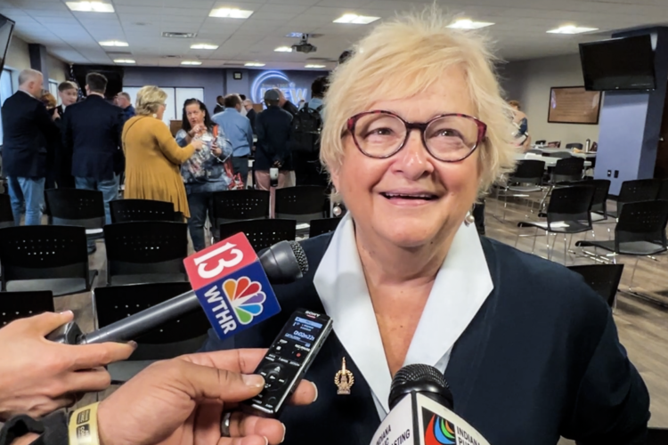 Karen Tallian is a White woman with blonde hair. She is wearing glasses and a black blazer over a light blue blouse.