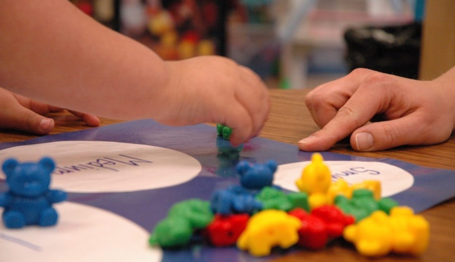 A tiny baby hand picks up a small green plastic bear from a pile of green, yellow, red and blue bears. A larger hand in the background points to a mat with appropriately-sized circles labeled small and medium.