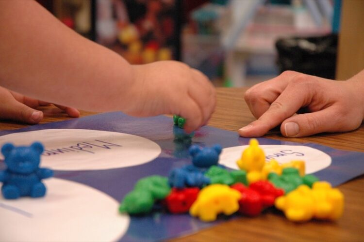 A tiny baby hand picks up a small green plastic bear from a pile of green, yellow, red and blue bears. A larger hand in the background points to a mat with appropriately-sized circles labeled small and medium.