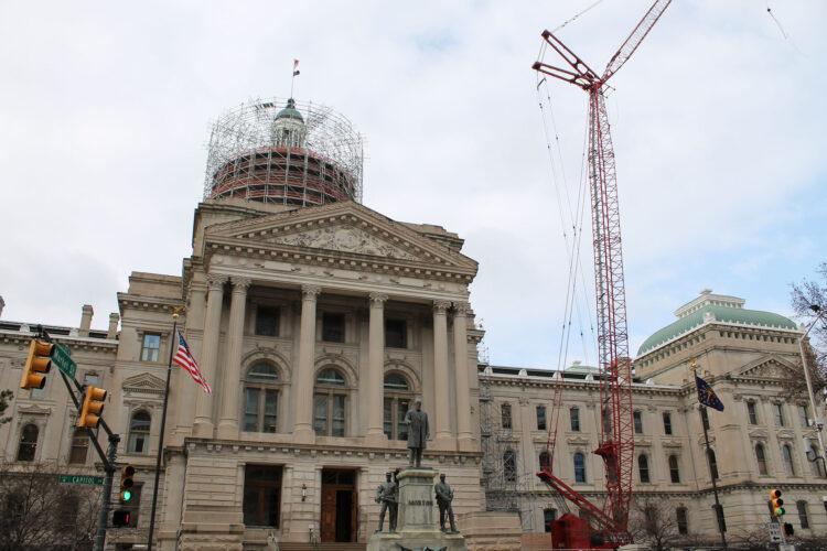 The Indiana Statehouse sits against a blue sky with a red construction crane visible to the right and scaffolding around the cupola.