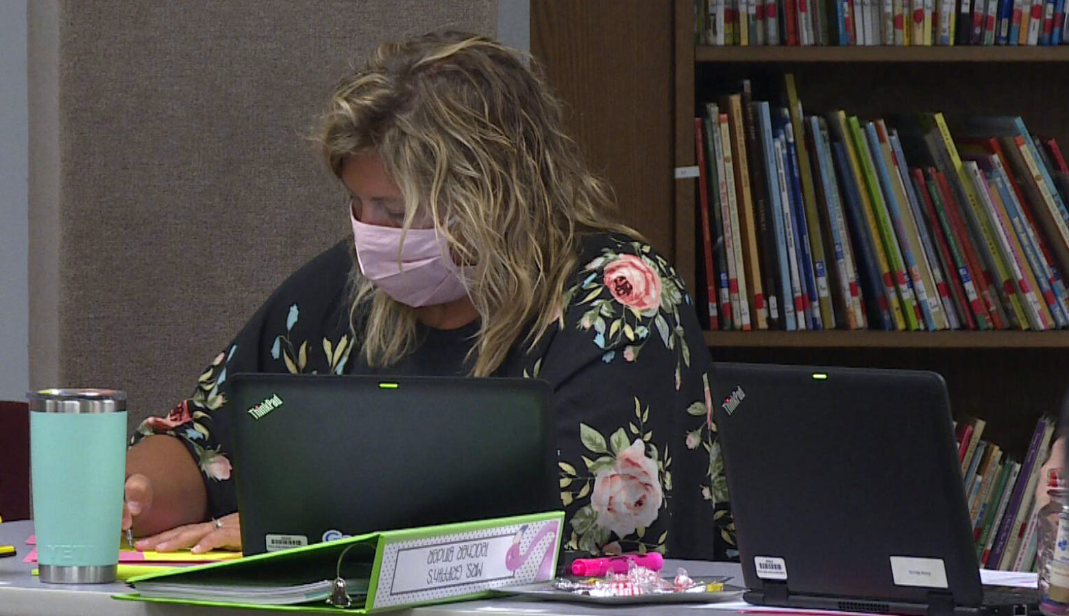 An educator wearing a fabric mask sits at her desk. She has several laptops surrounding her and a teal travel mug.