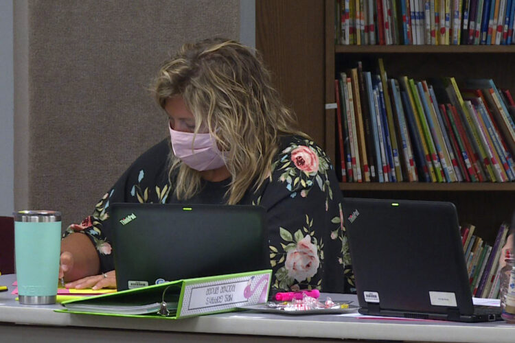 An educator wearing a fabric mask sits at her desk. She has several laptops surrounding her and a teal travel mug.