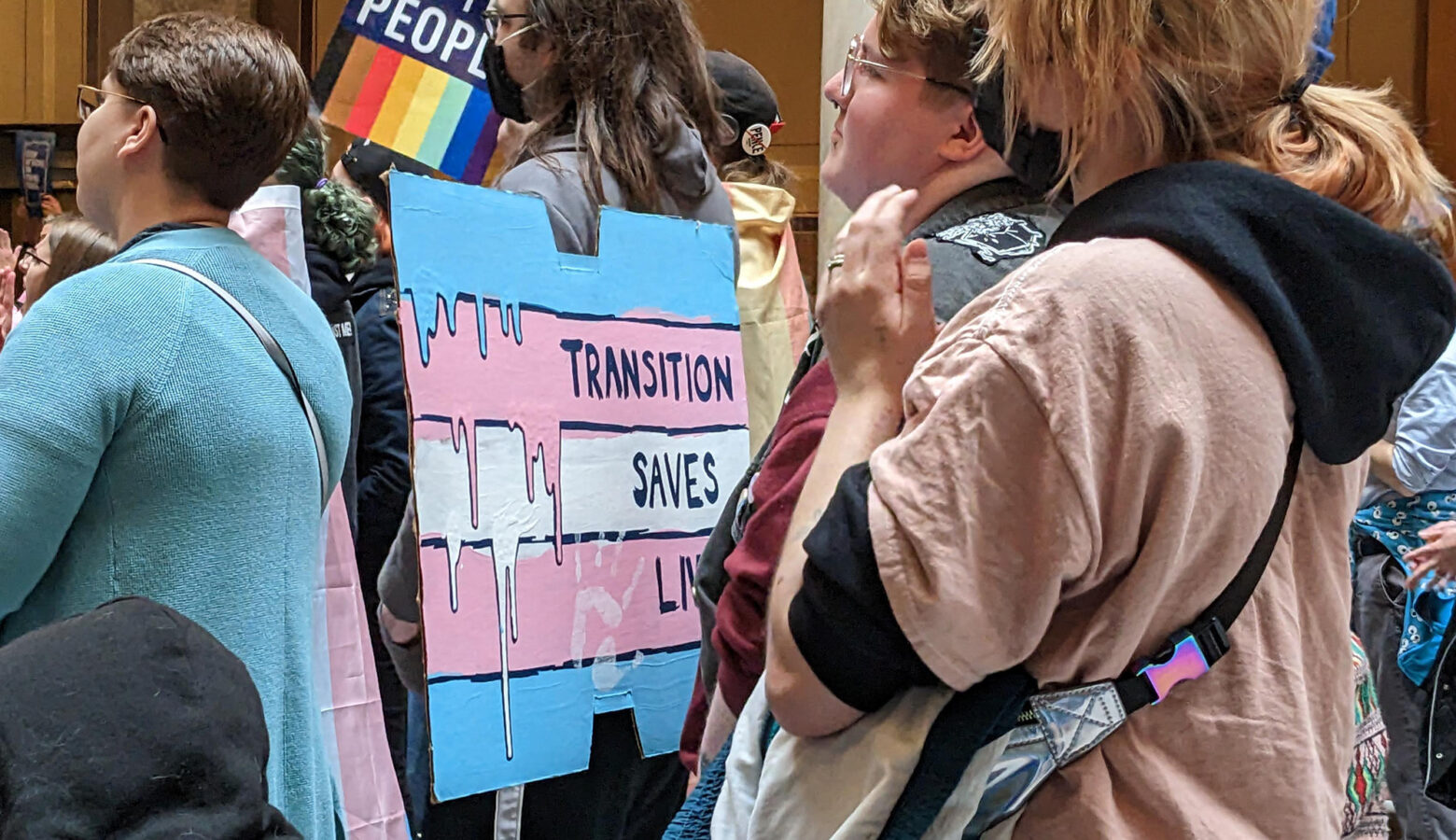 In a crowd at the Indiana Statehouse, a person holds a large protest sign that is painted to look like the transgender pride flag and has written on it "Transition saves lives."