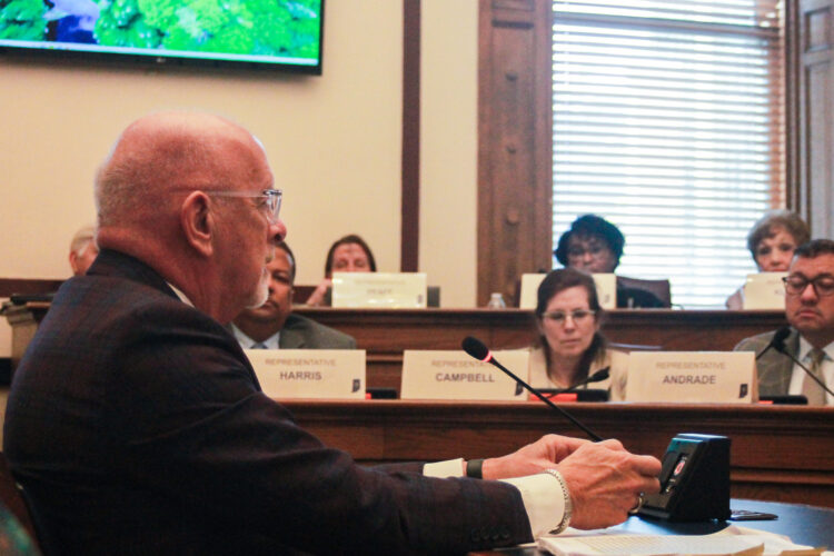 Travis Holdman sits at a table, speaking into a microphone. In the background, House committee members look on. Holdman is a White man, bald, with glasses and a white goatee. He is seen in profile, wearing a suit and tie.