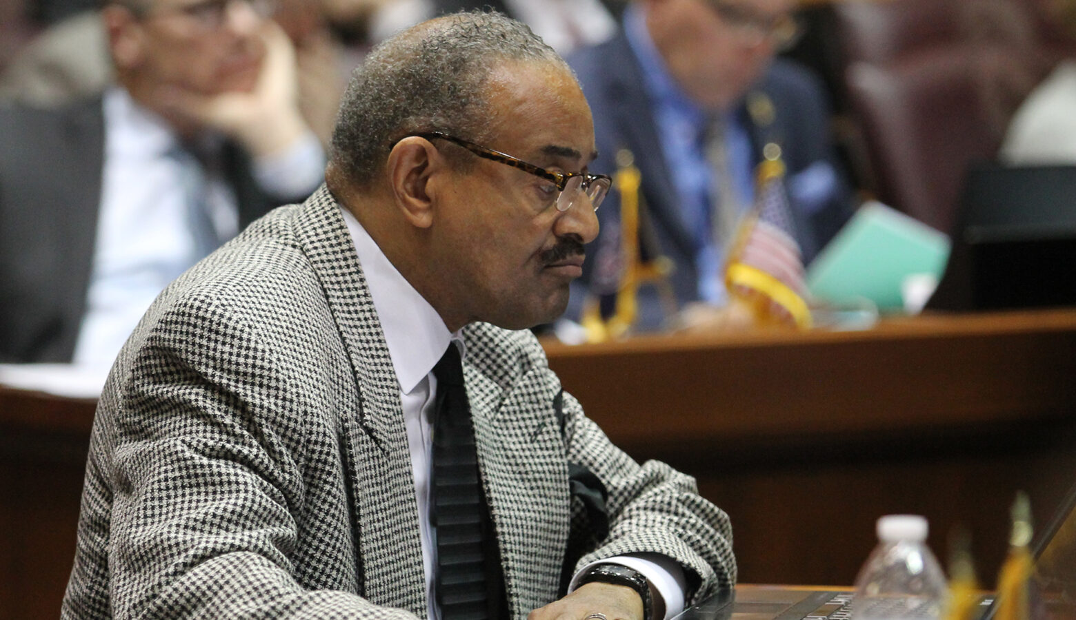 Vernon Smith wears a gray and black checkered suit and with glasses. He sits at a desk in the House chamber and looks forward. He is Black man with grey hair and a dark mustache.