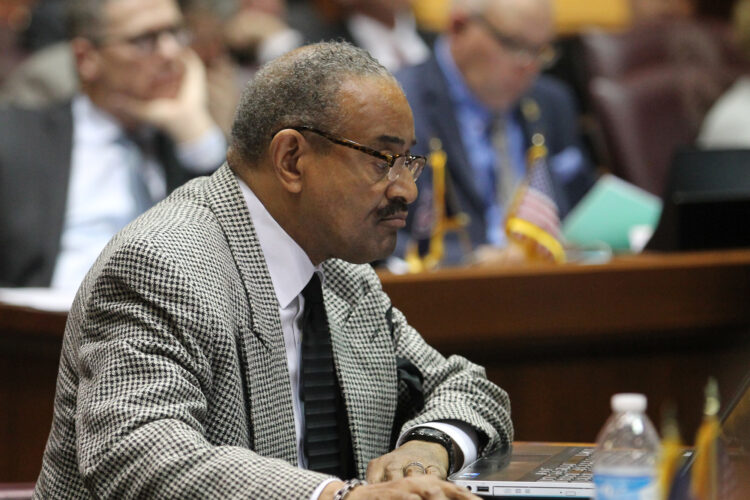 Vernon Smith wears a gray and black checkered suit and with glasses. He sits at a desk in the House chamber and looks forward. He is Black man with grey hair and a dark mustache.