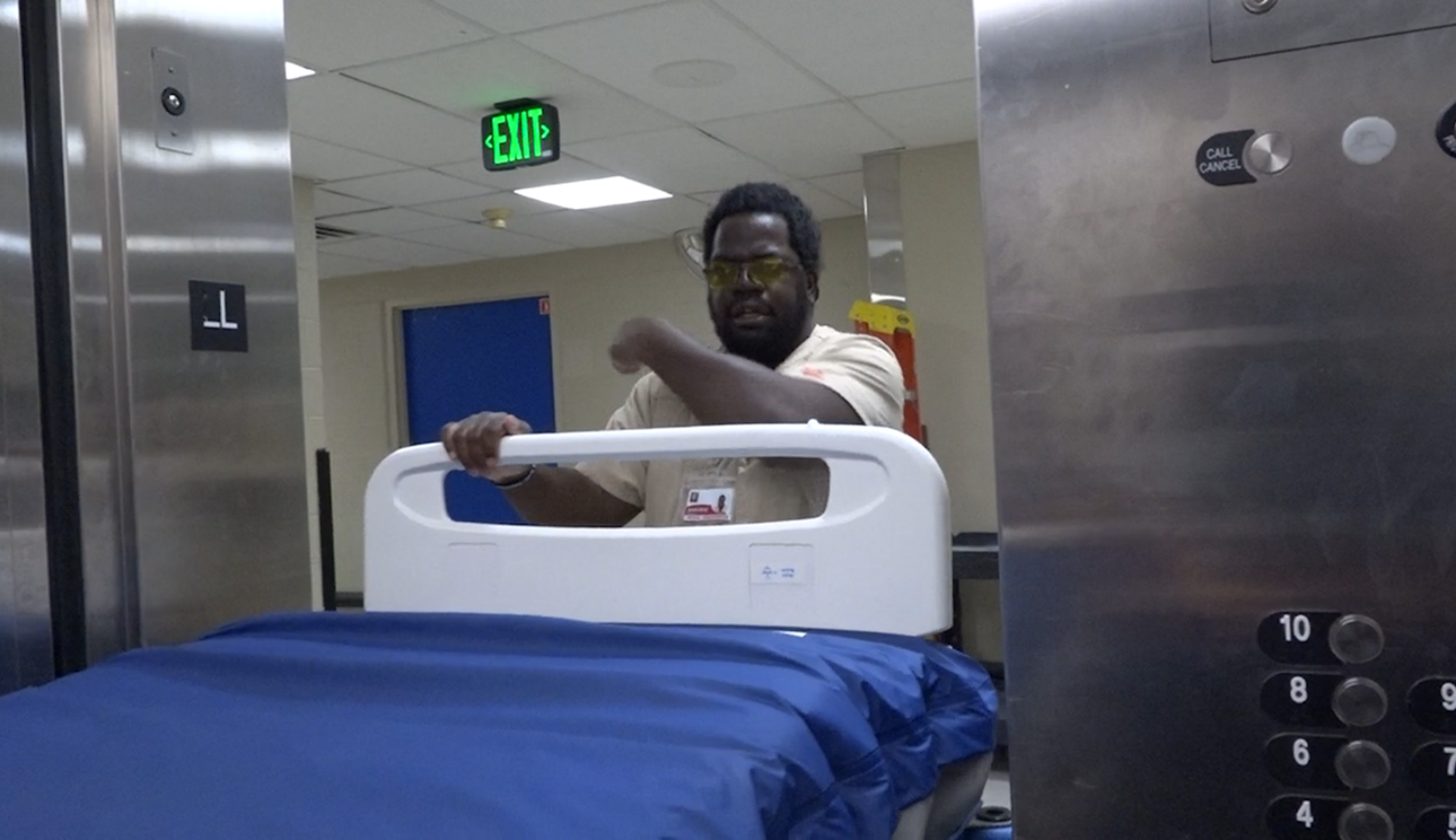 Jean-Rony Warner is moving a hospital bed to a storage room, pointing to the right side of him to indicate where he is going. Warner is a Black man with short hair and a beard. He is wearing tan scrubs and yellow-tinted glasses.