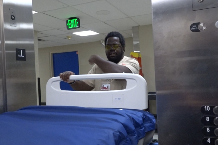 Jean-Rony Warner is moving a hospital bed to a storage room, pointing to the right side of him to indicate where he is going. Warner is a Black man with short hair and a beard. He is wearing tan scrubs and yellow-tinted glasses.