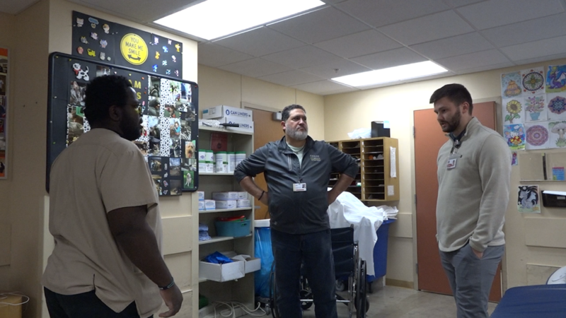Warner is talking to Jordan Starks. Starks is a White man with short hair and a beard. He is wearing blue jeans and an off-white pullover. Between them is EGTI’s Manager of Community Outreach, Daniel Gurulé. Gurulé is a White man with a buzz cut and a gray beard. He is wearing blue jeans and a gray pullover.