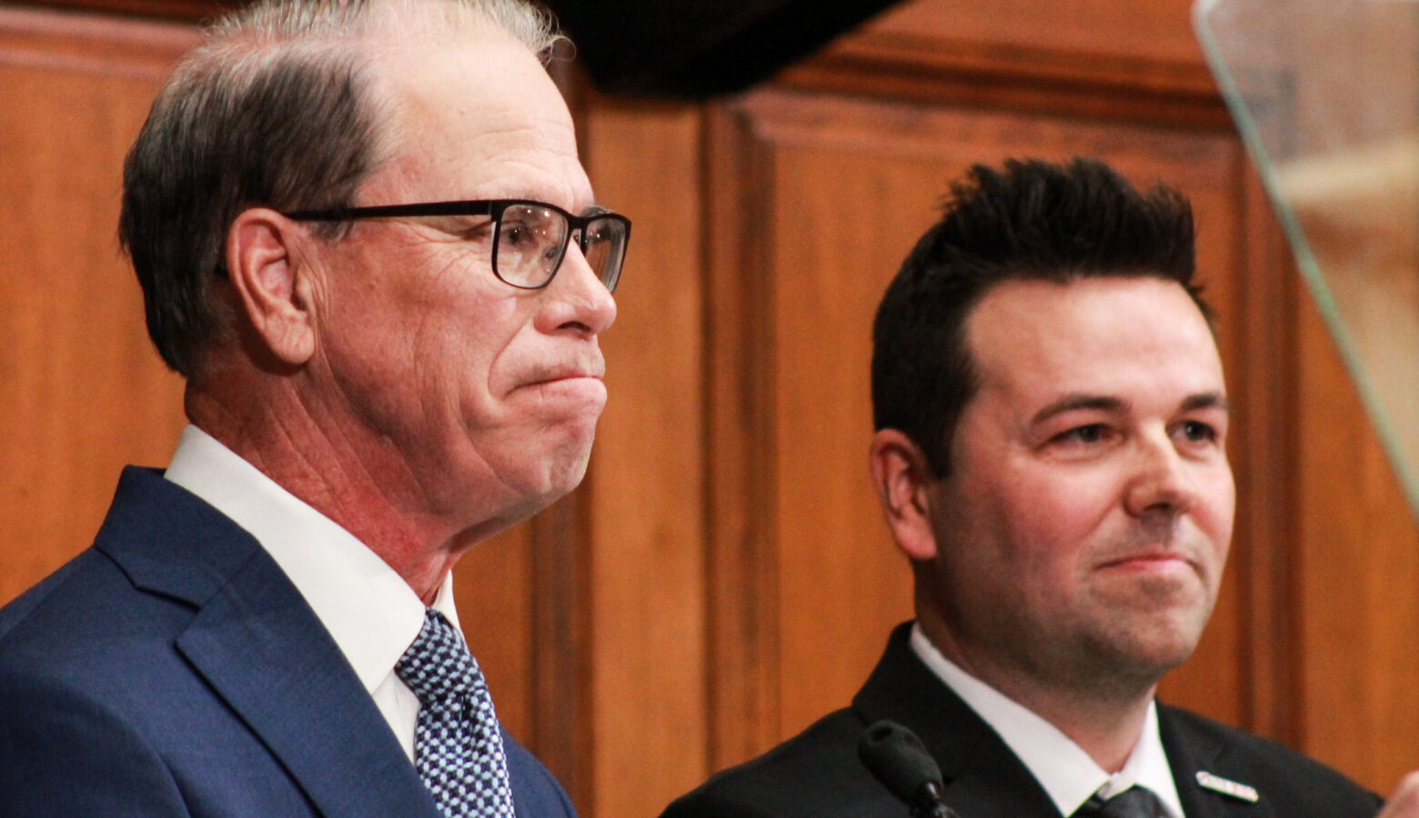 Mike Braun stands next to Micah Beckwith in the House chamber. Braun is a White man, balding with dark gray hair. He is wearing glasses and a blue suit. Beckwith is a White man with dark hair. He is wearing a black suit.