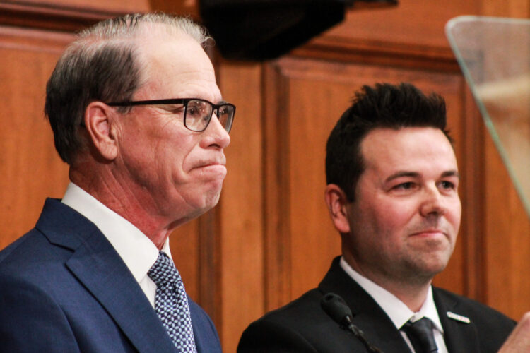 Mike Braun stands next to Micah Beckwith in the House chamber. Braun is a White man, balding with dark gray hair. He is wearing glasses and a blue suit. Beckwith is a White man with dark hair. He is wearing a black suit.