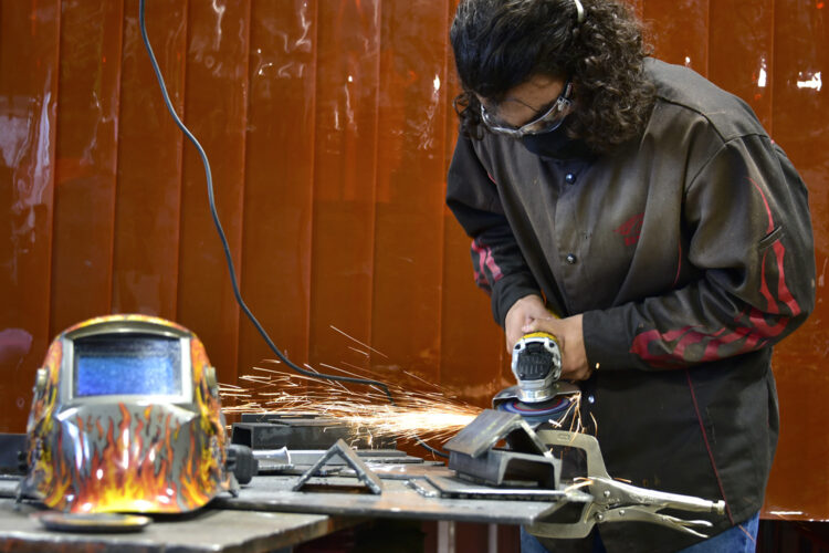 There is a student with a silver saw in his hand while he works on a project. There are sparks flying from the side of the saw. The student is wearing a black safety mask and safety glasses.