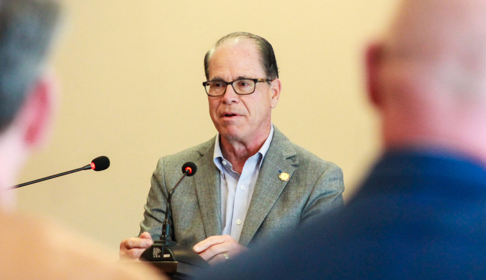 Mike Braun speaks at a lectern, framed between the backs of two people's heads in the foreground. Braun is a White man, balding with dark gray hair. He is wearing glasses and a sport coat over a dress shirt.