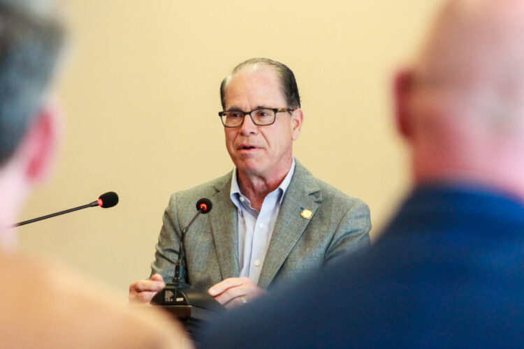 Mike Braun speaks at a lectern, framed between the backs of two people's heads in the foreground. Braun is a White man, balding with dark gray hair. He is wearing glasses and a sport coat over a dress shirt.