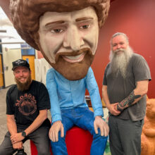 Two men sit on either side of a giant Bob Ross bobblehead. They're both smiling.