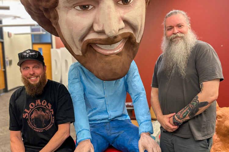 Two men sit on either side of a giant Bob Ross bobblehead. They're both smiling.