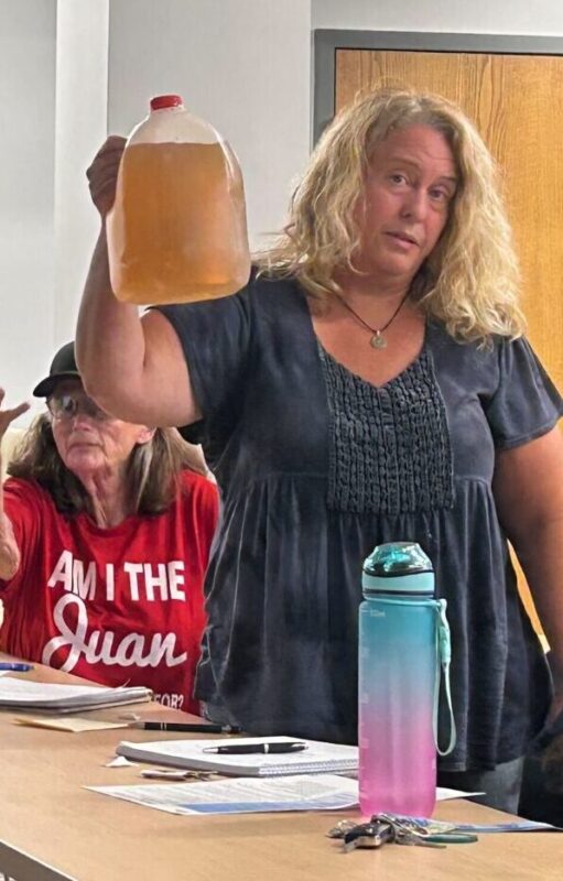 A women in a dark blue dress holds up a gallon jug of brown water.