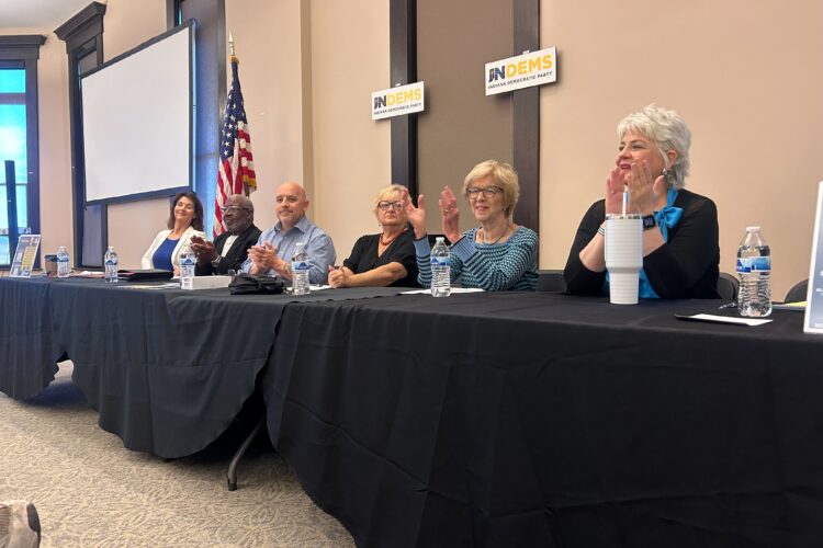 Multiple people sit behind a table with a black drape over it clapping.