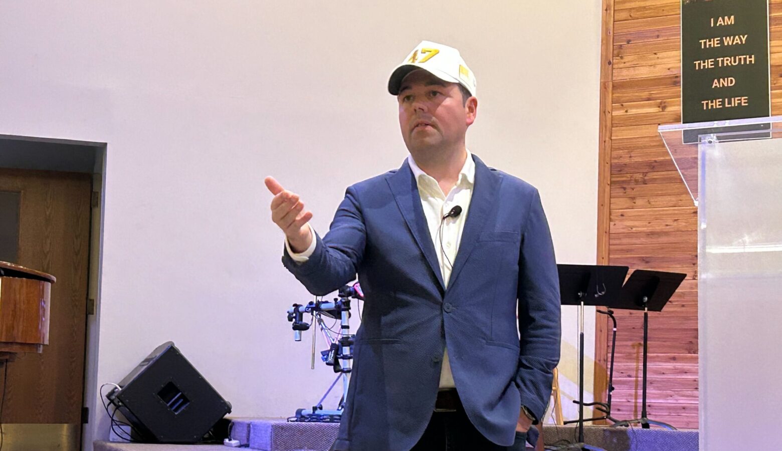 Lt. Gov. Micah Beckwith stands in the front of a room He is a White man in a blue suit with a white baseball cap.