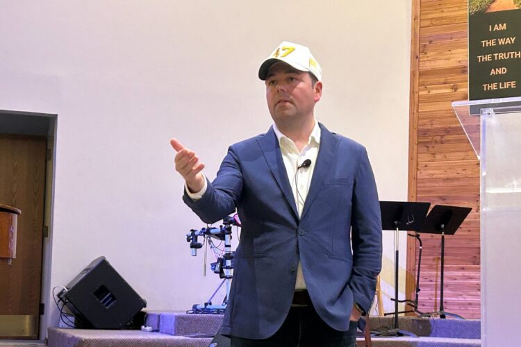 Lt. Gov. Micah Beckwith stands in the front of a room He is a White man in a blue suit with a white baseball cap.