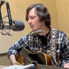 A man in a flannel sings into a microphone while playing guitar, his eyes are closed in concentration.