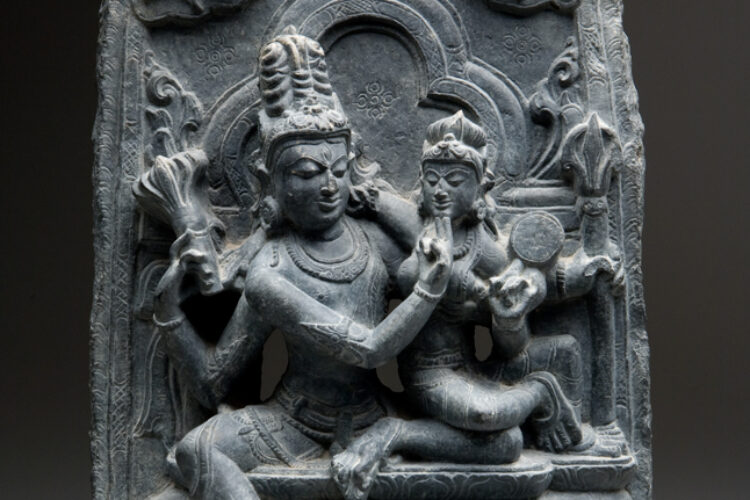 A grey stone sculpture of Uma Maheshvara, the divine couple Shiva and Parvati, shown in a loving embrace that reflects spiritual unity and cosmic balance.
