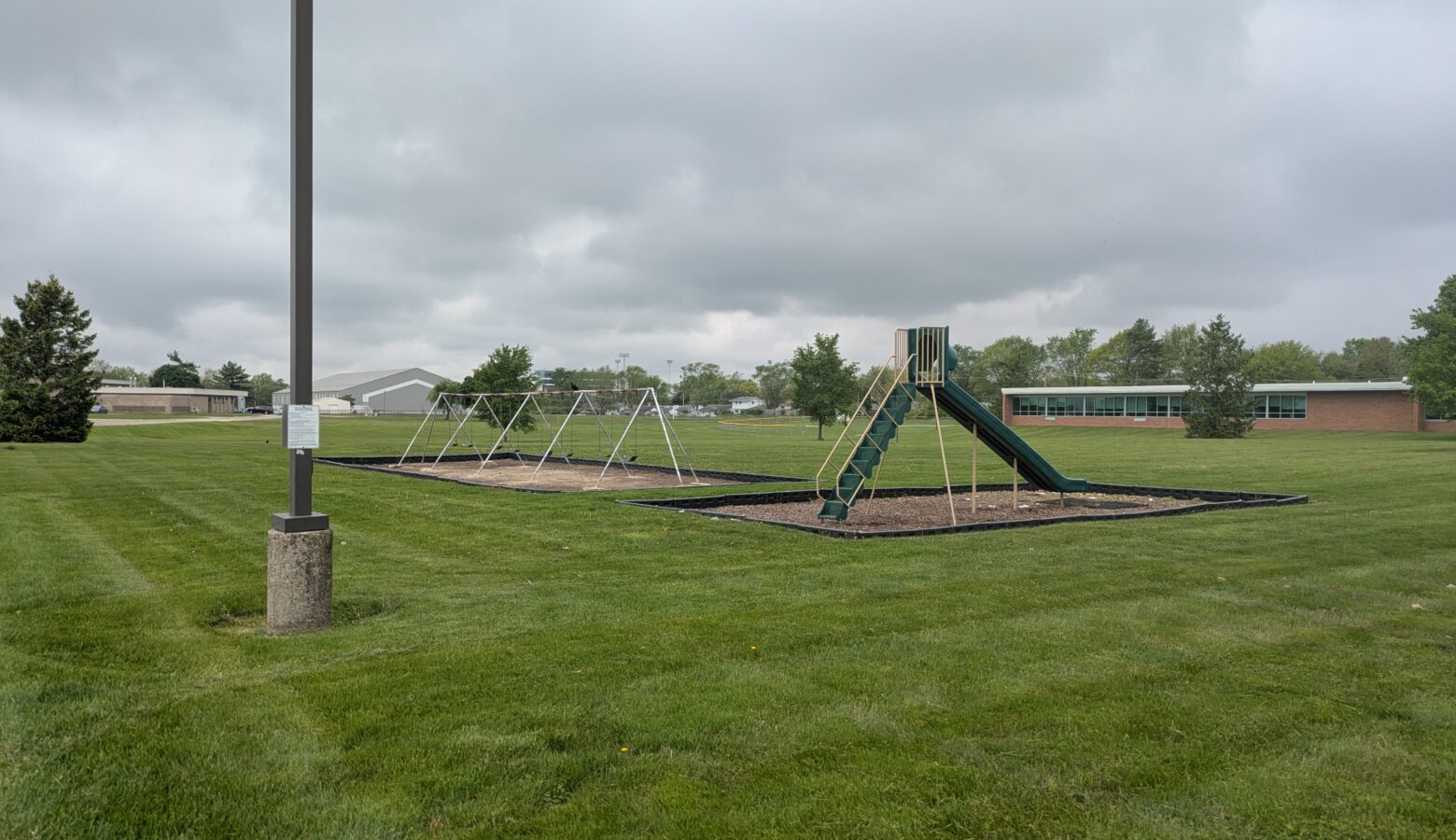 A metal A-frame full of swings and a green plastic enclosed slide sits on a long green lawn outside a brick building.
