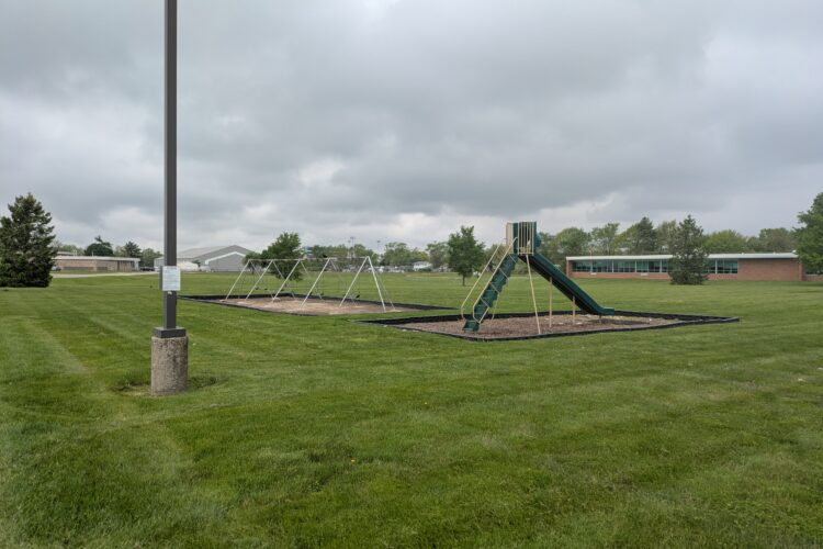 A metal A-frame full of swings and a green plastic enclosed slide sits on a long green lawn outside a brick building.