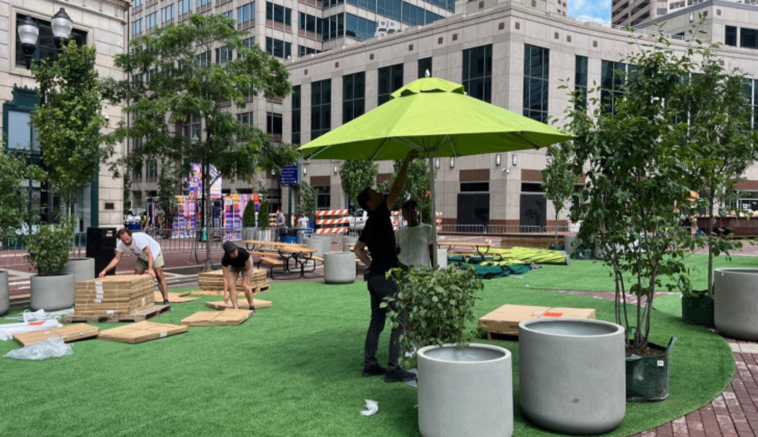Workers on Monument Circle are placing lawn furniture and large, green shade umbrellas on artificial grass to resemble a public park. Small potted trees are also in place, amid downtown Indianapolis buildings.
