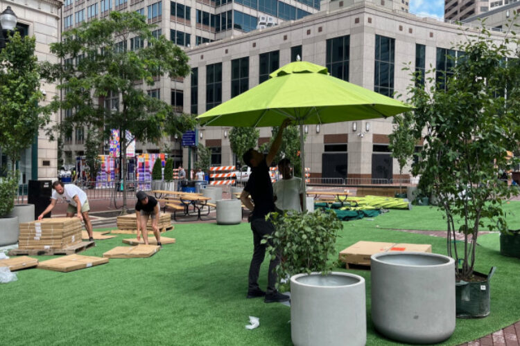 Workers on Monument Circle are placing lawn furniture and large, green shade umbrellas on artificial grass to resemble a public park. Small potted trees are also in place, amid downtown Indianapolis buildings.