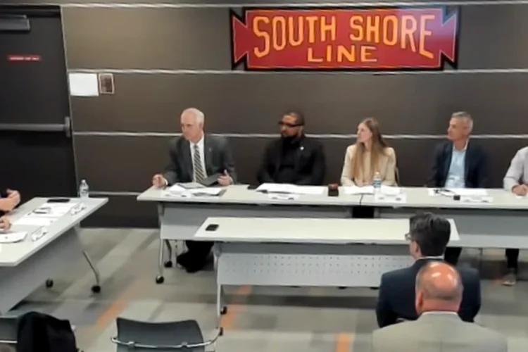 NICTD board members seated at two tables in an L configuration. The classic South Shore Line logo is attached to a wall. The logo is red with yellow letters and a black border.