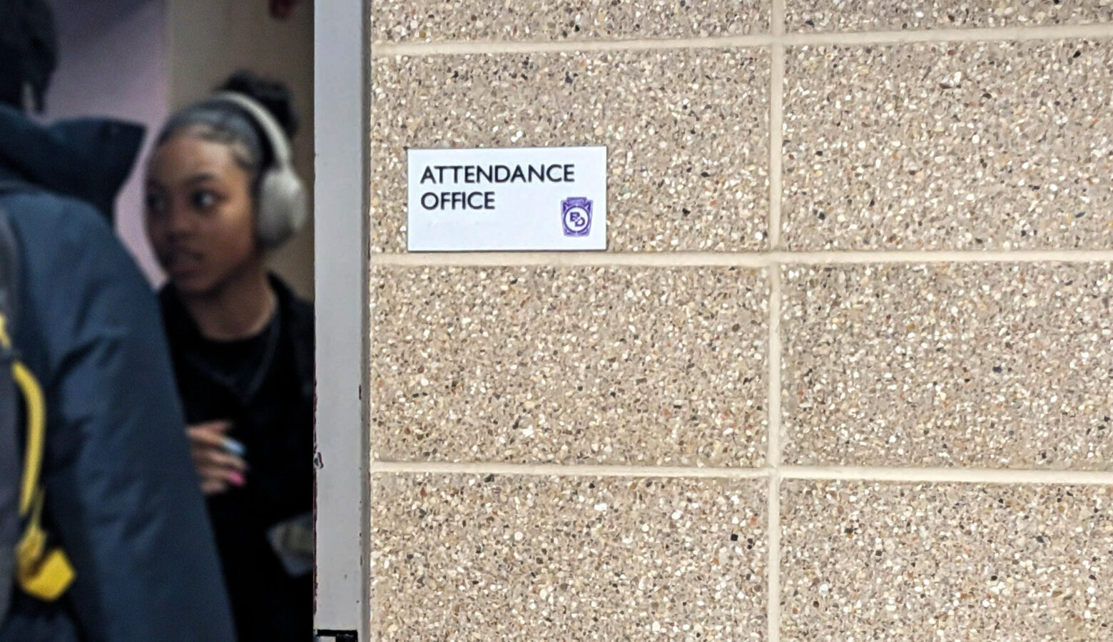 A small silver plaque that says "attendance office" is posted on a tan brick wall and students are visible beyond the edge of the doorway