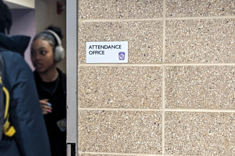 A small silver plaque that says "attendance office" is posted on a tan brick wall and students are visible beyond the edge of the doorway