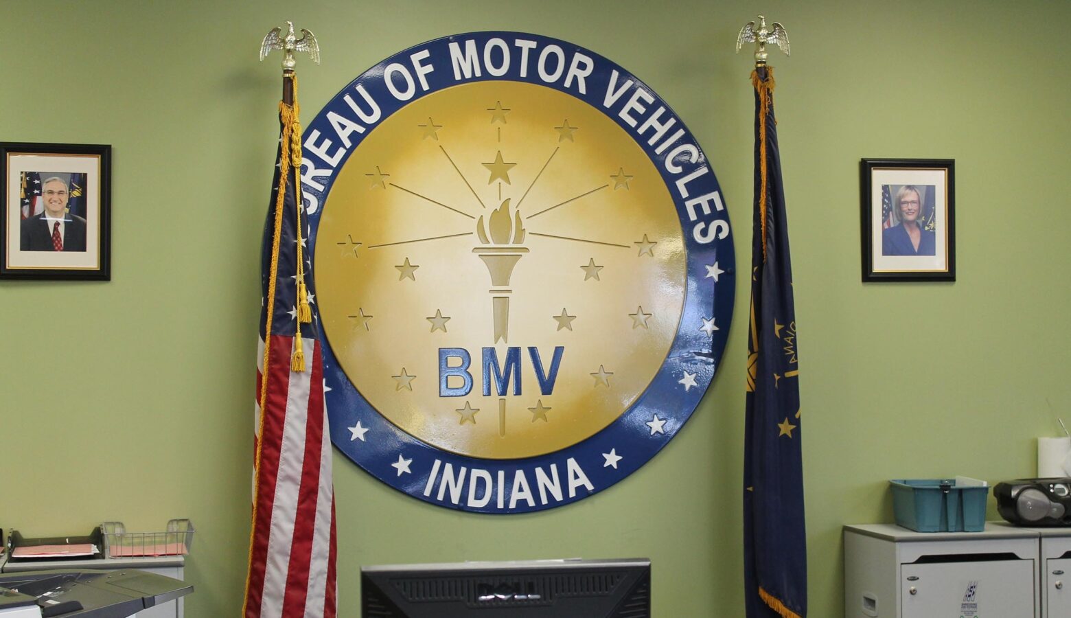 The Indiana BMV seal hangs on a green wall, surrounded by the American and Indiana flags.