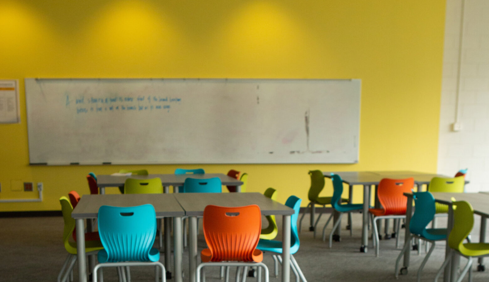 An empty classroom with unoccupied tables and chairs with a white dry erase board attached to a yellow wall.